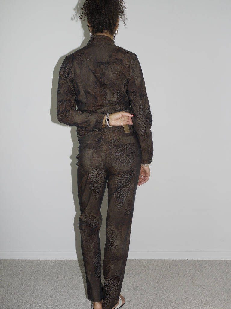 Model wearing a matching vintage brown set with subtle floral patterns, consisting of a long-sleeve button-up shirt and straight-leg pants. Styled barefoot in flip-flops, photographed against a plain white wall. The look has a 90s minimalist vibe with a soft utilitarian edge.