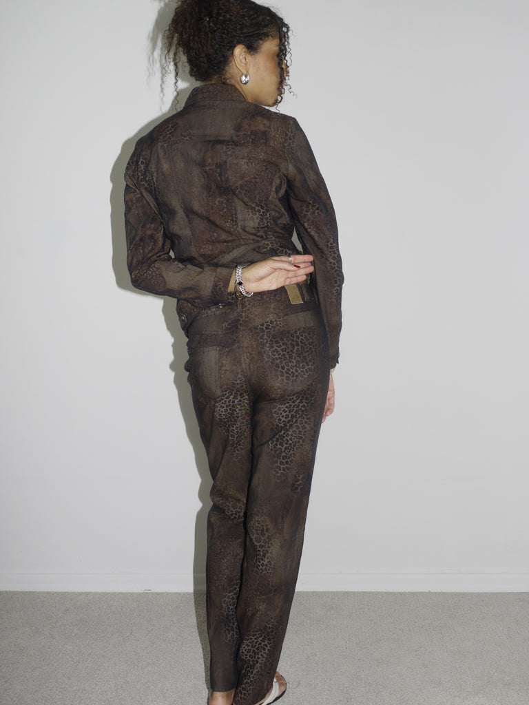 Model wearing a matching vintage brown set with subtle floral patterns, consisting of a long-sleeve button-up shirt and straight-leg pants. Styled barefoot in flip-flops, photographed against a plain white wall. The look has a 90s minimalist vibe with a soft utilitarian edge.