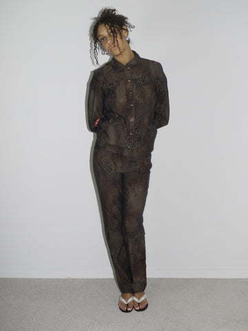 Model wearing a matching vintage brown set with subtle floral patterns, consisting of a long-sleeve button-up shirt and straight-leg pants. Styled barefoot in flip-flops, photographed against a plain white wall. The look has a 90s minimalist vibe with a soft utilitarian edge.