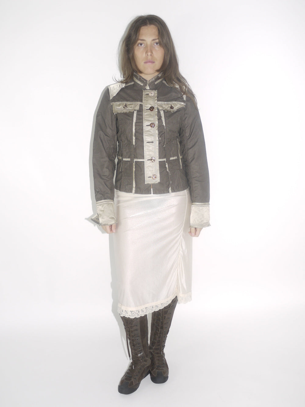 Model wearing a structured brown Guess jacket with beige stitching and lace-trimmed cuffs, paired with a cream satin midi skirt and knee-high brown boots. The look blends vintage western tailoring with soft romantic details