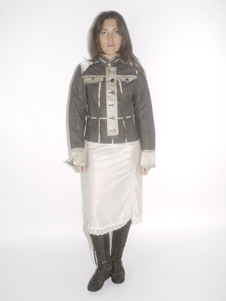 Model wearing a structured brown Guess jacket with beige stitching and lace-trimmed cuffs, paired with a cream satin midi skirt and knee-high brown boots. The look blends vintage western tailoring with soft romantic details