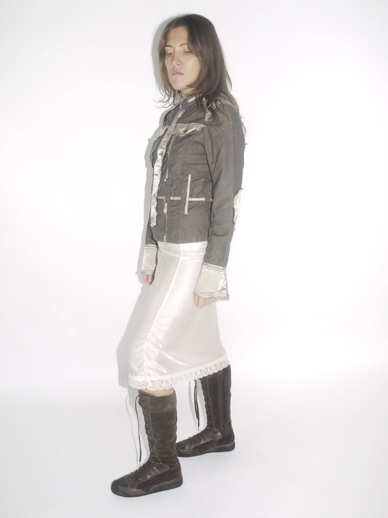 Model wearing a structured brown Guess jacket with beige stitching and lace-trimmed cuffs, paired with a cream satin midi skirt and knee-high brown boots. The look blends vintage western tailoring with soft romantic details