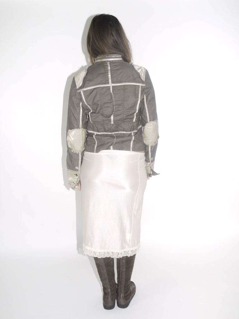 Model wearing a structured brown Guess jacket with beige stitching and lace-trimmed cuffs, paired with a cream satin midi skirt and knee-high brown boots. The look blends vintage western tailoring with soft romantic details