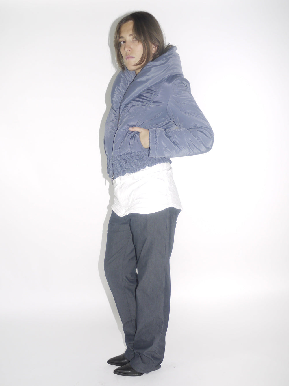 Model wearing a cropped metallic blue Charactère puffer jacket with a high funnel collar and textured waistband, layered over a white top and paired with dark straight-leg trousers and pointed black boots. A bold structured winter silhouette with a futuristic edge.
