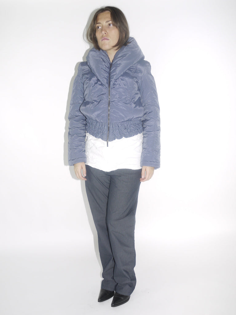 Model wearing a cropped metallic blue Charactère puffer jacket with a high funnel collar and textured waistband, layered over a white top and paired with dark straight-leg trousers and pointed black boots. A bold structured winter silhouette with a futuristic edge.