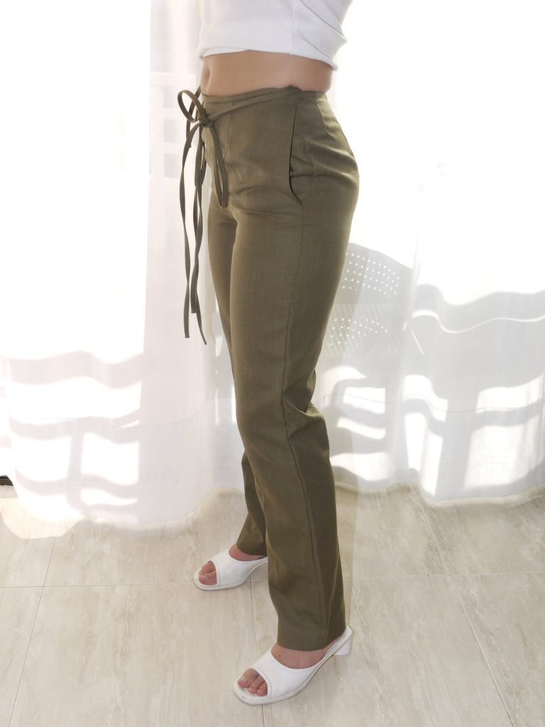 Model wearing a long-sleeve black top with asymmetrical seams, paired with iconic olive green Maison Margiela pants featuring deconstructed strap detailing hanging from the waistband. Styled with black-and-white printed heels for a conceptual, avant-garde look. Shot against a plain white background.