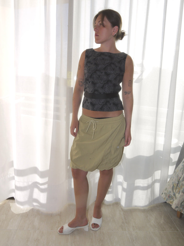 Model wearing a sleeveless fitted top with black floral jacquard pattern, paired with a low-rise beige mini skirt featuring drawstring details and a sporty silhouette. Barefoot styling gives it a casual, lived-in feel. Shot against a plain white wall.