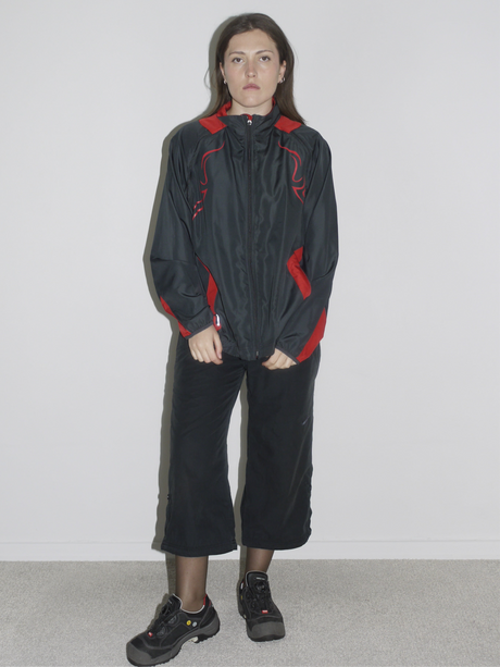 Model wearing a black zip-up windbreaker with red tribal-style racing details, paired with black cropped cargo pants and chunky black sneakers. The look blends Y2K motorsport energy with urban utilitarianism — reminiscent of early 2000s Puma, McQueen x Puma, and underground techwear.