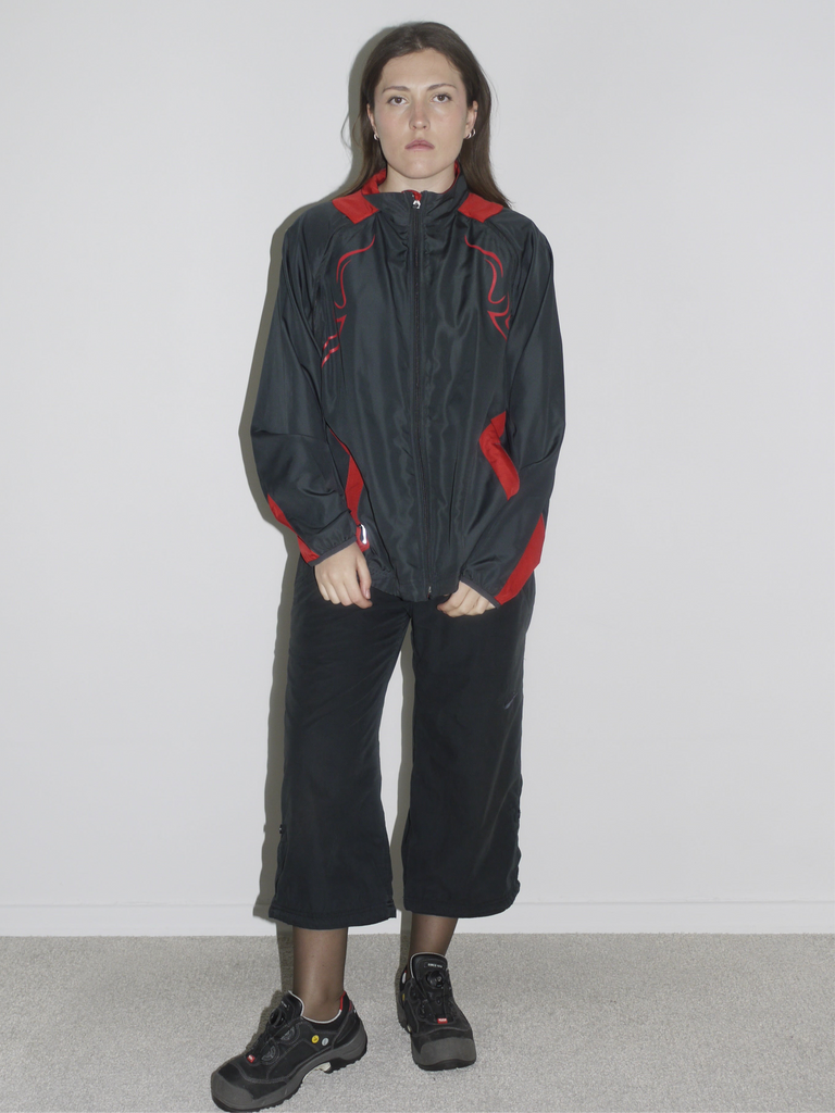 Model wearing a black zip-up windbreaker with red tribal-style racing details, paired with black cropped cargo pants and chunky black sneakers. The look blends Y2K motorsport energy with urban utilitarianism — reminiscent of early 2000s Puma, McQueen x Puma, and underground techwear.