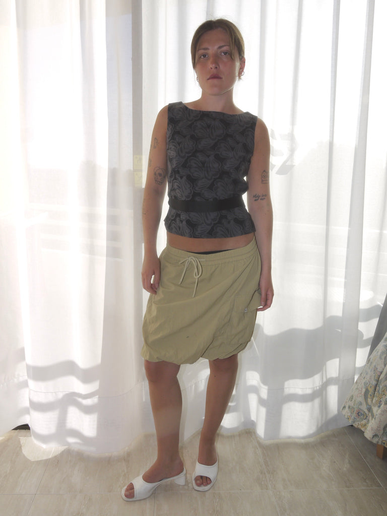 Model wearing a sleeveless fitted top with black floral jacquard pattern, paired with a low-rise beige mini skirt featuring drawstring details and a sporty silhouette. Barefoot styling gives it a casual, lived-in feel. Shot against a plain white wall.