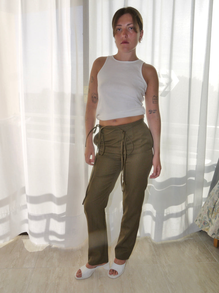 Model wearing a long-sleeve black top with asymmetrical seams, paired with iconic olive green Maison Margiela pants featuring deconstructed strap detailing hanging from the waistband. Styled with black-and-white printed heels for a conceptual, avant-garde look. Shot against a plain white background.