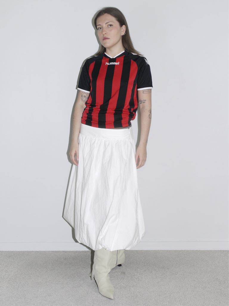 Model wearing a vintage Hummel football jersey in red and black stripes, styled with a voluminous white balloon skirt and off-white cowboy boots. A sporty-feminine mix with early 2000s energy — like AC Milan meets Paloma Wool.