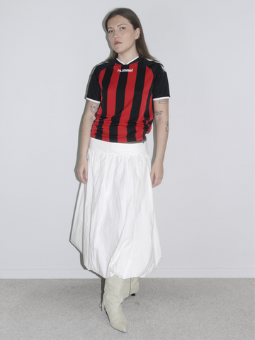 Model wearing a vintage Hummel football jersey in red and black stripes, styled with a voluminous white balloon skirt and off-white cowboy boots. A sporty-feminine mix with early 2000s energy — like AC Milan meets Paloma Wool.