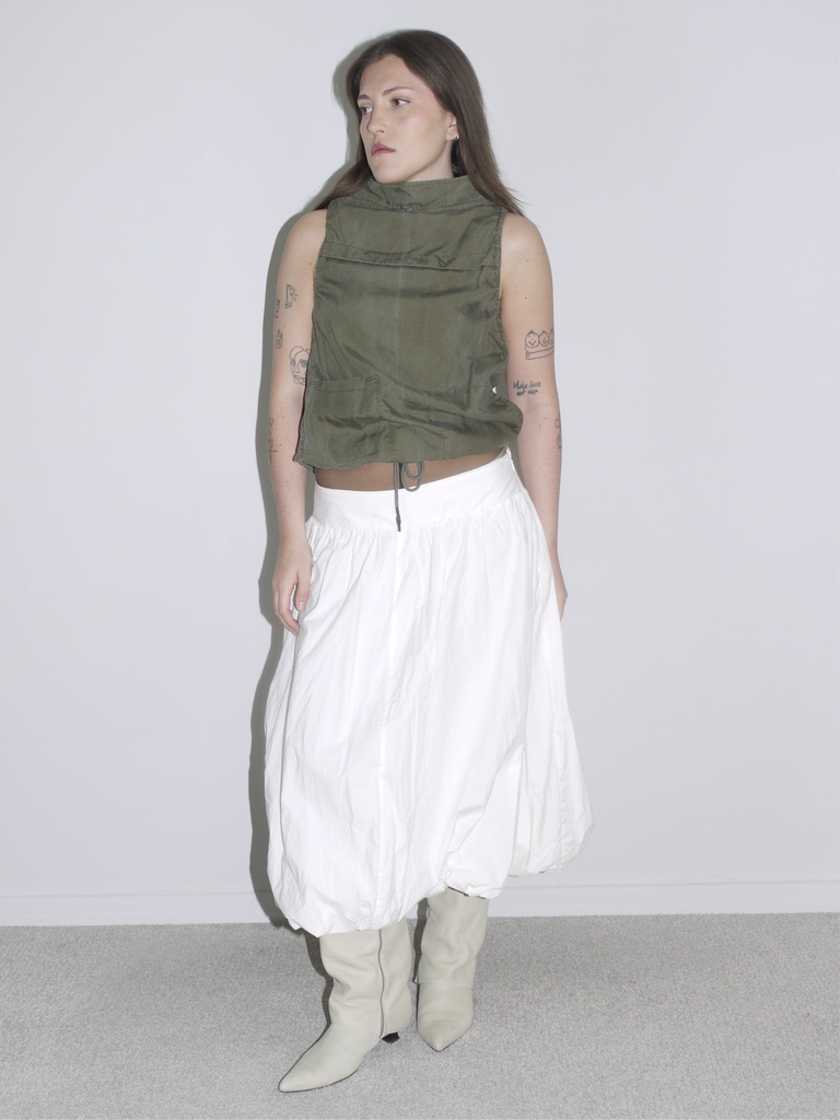 Minimalist desert-inspired outfit with an olive green sleeveless high-neck top featuring drawstring and flap pockets, styled with a voluminous white balloon skirt and cream western boots.