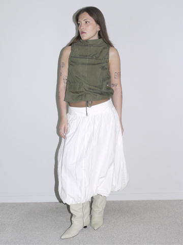 Minimalist desert-inspired outfit with an olive green sleeveless high-neck top featuring drawstring and flap pockets, styled with a voluminous white balloon skirt and cream western boots.