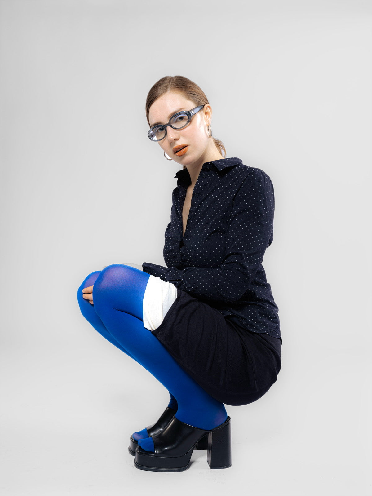 Model wearing a black sheer blouse with textured dots, styled with wide brown trousers and electric blue tights – edgy, archive-inspired look with platform heels and rectangular sunglasses.