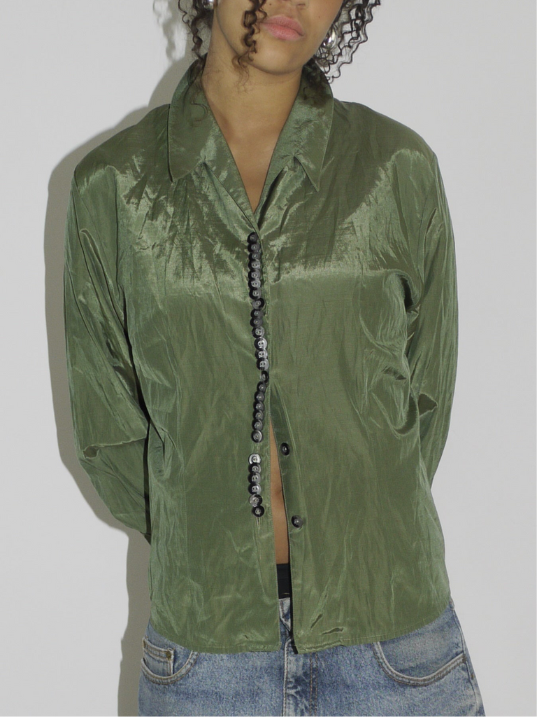 Model wearing a crinkled olive green button-up shirt, left slightly unbuttoned to reveal a black waistband, paired with loose-fitting vintage denim Bermuda shorts. Styled with pointed black-and-white printed heels for a contrast of grunge and glam. Shot against a plain white background.