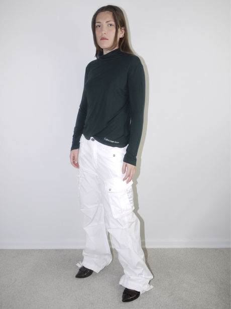Model wearing a fitted forest green Calvin Klein turtleneck top with logo detail at the hem, paired with wide white cargo pants and pointed black flats — sleek and minimal Y2K streetwear look, photographed against clean white wall.