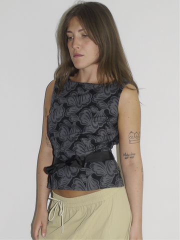 Model wearing a sleeveless fitted top with black floral jacquard pattern, paired with a low-rise beige mini skirt featuring drawstring details and a sporty silhouette. Barefoot styling gives it a casual, lived-in feel. Shot against a plain white wall.