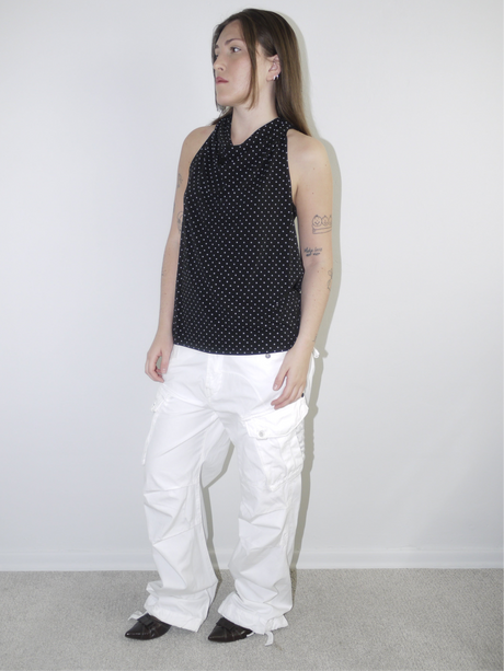 Model wearing a sleeveless black halterneck top with fine white polka dots, styled with white wide-leg cargo trousers and pointed flats — playful yet clean Y2K-inspired look, photographed against plain white background.