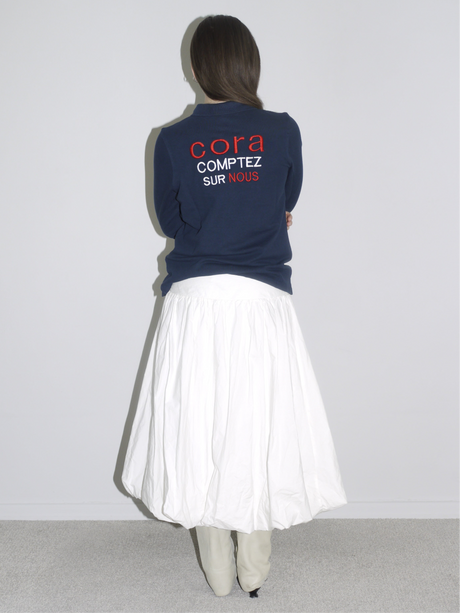 Back view of a navy blue French logo sweatshirt reading “Cora – Comptez sur nous,” styled with a voluminous white balloon skirt and pointed cream boots. A casual meets editorial streetwear look with vintage flair.
