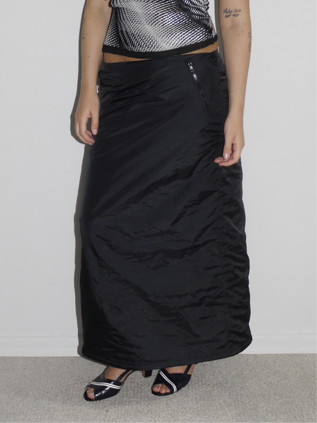 Model wearing a sleeveless metallic black-and-silver optical print top with a fitted silhouette, paired with a black low-rise satin-effect maxi skirt. Styled with open-toe slingback heels for a sleek, futuristic evening look. Shot against a plain white wall