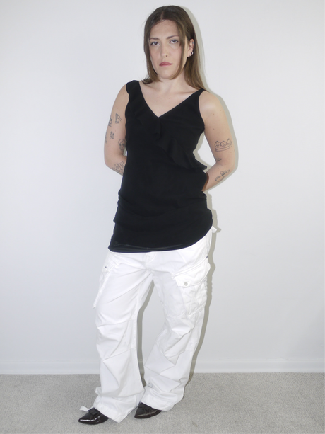 Model wearing a black asymmetrical waterfall draped top styled as a mini dress, paired with white utility cargo pants and pointed black heels — deconstructed Y2K-inspired layering look with soft volume and streetwear edge, shot against plain white wall.