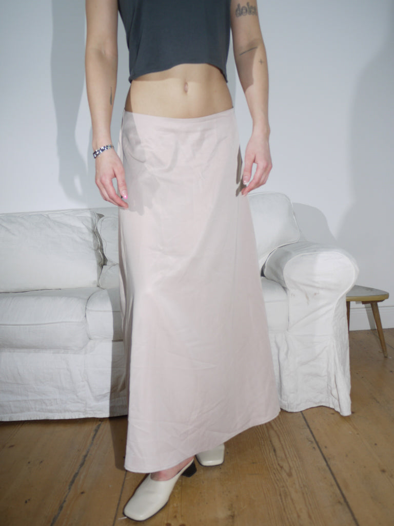 Model wearing a soft beige low-rise maxi skirt with a fluid, slightly flared silhouette. Styled with a cropped black top and cream leather mules for an understated, modern look.