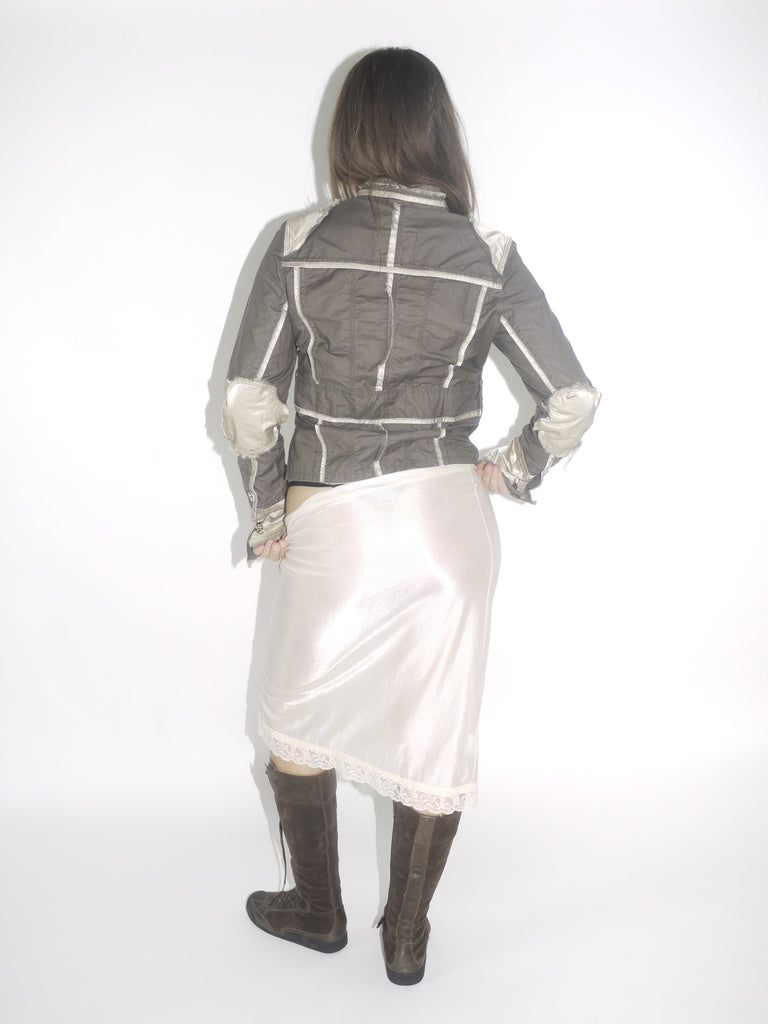 Model wearing a structured brown Guess jacket with beige stitching and lace-trimmed cuffs, paired with a cream satin midi skirt and knee-high brown boots. The look blends vintage western tailoring with soft romantic details